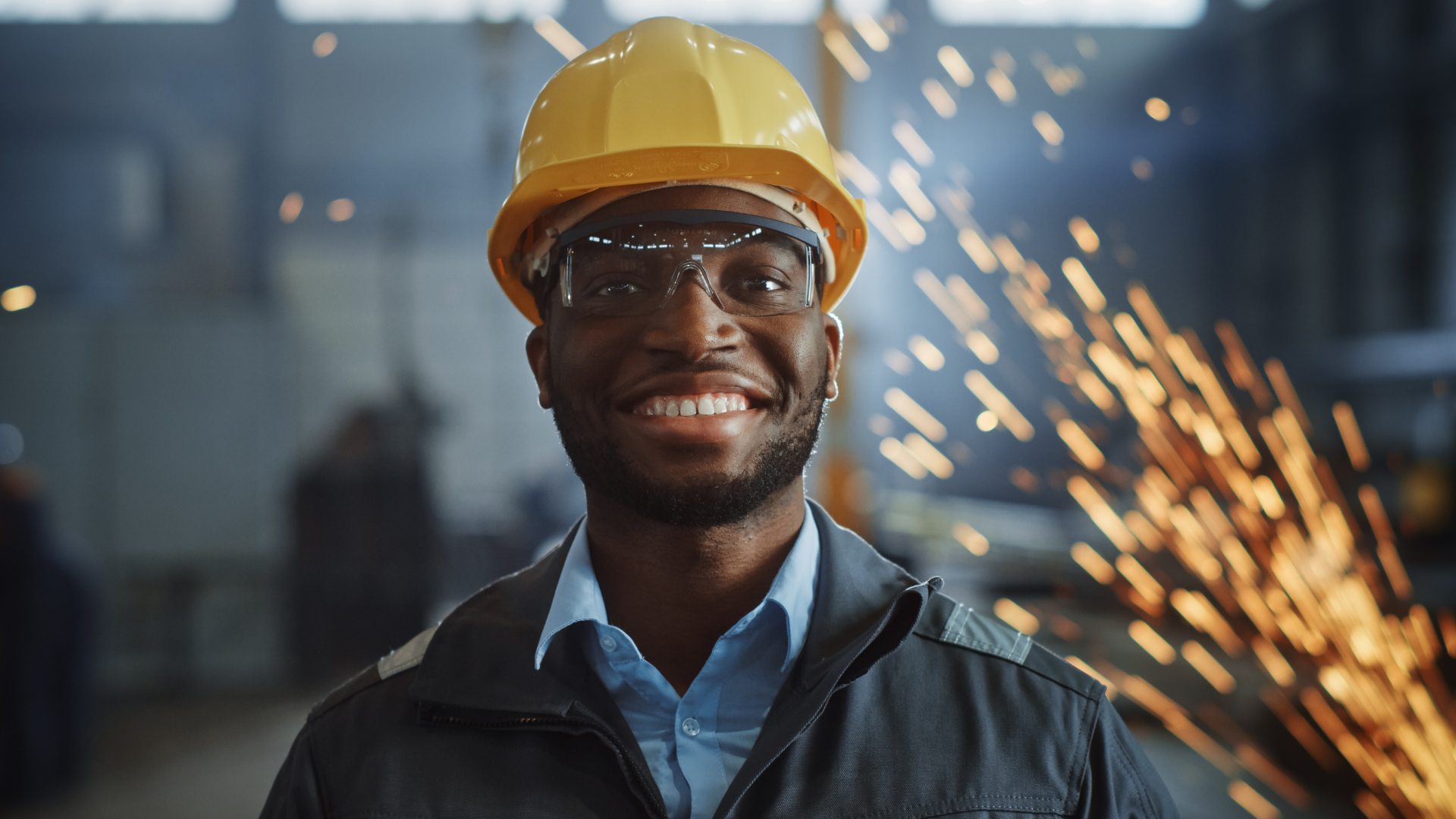 Happy Professional Heavy Industry Engineer/Worker Wearing Uniform, Glasses and Hard Hat in a Steel Factory. Smiling African American Industrial Specialist Standing in a Metal Construction Manufacture